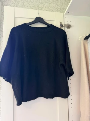 Svart oversized stickad kortärmad tröja - En svart, oversized stickad tröja med rund halsringning och kortare ärmar. Tröjan har en enkel design utan synliga detaljer eller mönster och är gjord i ett mjukt, lite luddigt material. Perfekt för en avslappnad och trendig look.