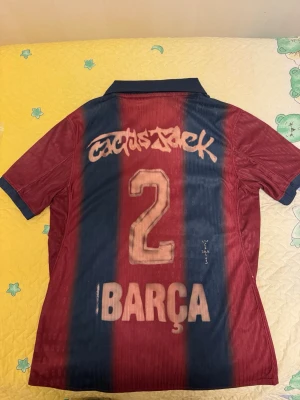 Barcelona Retro Travis Scott  -  Travis Scott x Barcelona player version. Tröjan har V-ringning, krage och korta ärmar. Perfekt för dig som älskar fotboll och vill visa ditt stöd för Barça.