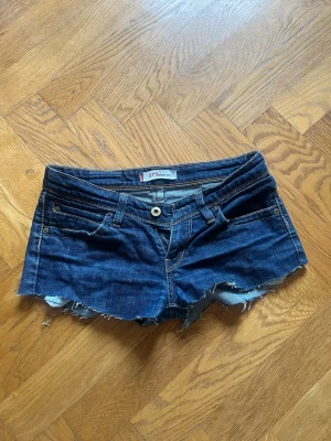 Levi's blå jeansshorts med rå kant - Säljer ett par klassiska blå jeansshorts från Levi's med rå, fransig kant och femficksmodell. Shortsen har låg midja och slitna detaljer för en avslappnad look. Perfekta till sommaren och lätta att matcha med allt. Har klippt dem till shorts själv. Står ingen storlek men passar folk som har xs-s.