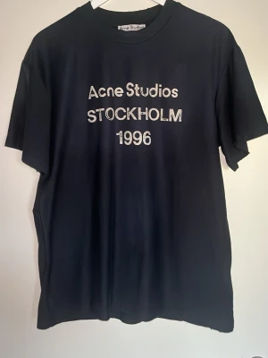 Svart Acne Studios t-shirt - Snygg svart t-shirt från Acne Studios med tryck 'Acne Studios STOCKHOLM 1996' på bröstet. Klassisk passform med rund hals och korta ärmar. Perfekt basplagg för en clean och stilren look.