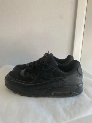 Nike Air Max 90 helsvarta sneakers - Säljer ett par helsvarta Nike Air Max 90 sneakers med klassisk siluett. Skorna har ovandel i en mix av läder och mesh, samt den ikoniska Air Max-dämpningen i sulan. Snygga detaljer och diskret Nike-logga på sidan. Perfekta för dig som gillar stilrena och tidlösa sneakers.