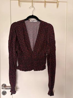 Blommig croppad blus från NA-KD - Trendig blommig blus från NA-KD med svart bas och små röda blommor. Djup v-ringning, croppad passform och långa ärmar med smock i midjan och vid ärmslut. Perfekt för dig som gillar en feminin och modern stil.