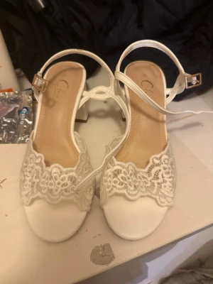 Vita klackar - Säljer ett par vita klackar med spetsdetaljer och öppen tå. Skorna har en elegant rem med guldfärgat spänne och låg klack. Perfekta för dig som vill ha en romantisk och feminin look. Materialet är en mix av tyg och spets. Helt oanvända, endast testade