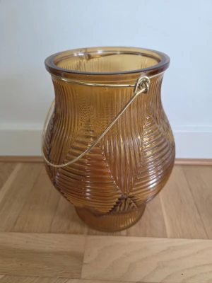 Ljuslykta bärnstensfärgat glas från Mio,  25 cm  - Mio ljuslykta  i bärnstensfärgat glas med räfflat mönster och guldfärgat metallhandtag. Perfekt som dekoration eller för att skapa mysig stämning med ett värmeljus. Glaset har en rundad form och fina detaljer.