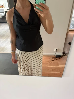 Svart halterneck topp med öppen rygg - Snygg svart halterneck topp med djup urringning och öppen rygg. Toppen knyts i nacken och har en mjuk, något glansig yta som ger en elegant känsla. Perfekt att styla med kjol eller jeans för en trendig look.