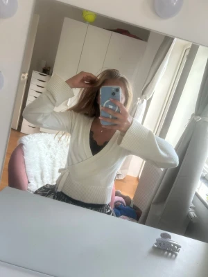 Vit omlottstickad kofta - Säljer en vit stickad omlottkofta med knytband i midjan och bred ribbad kant nedtill. Koftan har långa ärmar och en mjuk, skön känsla. Koftan är lite noprig men inget som syns tydligt vid användning. Den är i storlek 158/164 som motsvarar xs/ s med långa ärmar och lite kortare nedtill. 