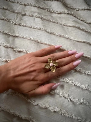 Blomring i rostfritt stål - Unik ring i guldfärgad metall formad som en blomma med fyra blad och en ljusrosa prick i mitten. 💕Justerbar storlek men ganska svår att böja så skulle utgå från storleken som står på bilden!🫰 Rostfritt stål och använd fåtal gånger! Hör av er vid frågor☀️
