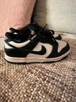 Nike Dunk Low svartvita sneakers - Säljer ett par Nike Dunk Low sneakers i svart och vitt. Klassisk låg modell med svart swoosh, vita paneler och svarta detaljer. Skorna har snörning och perforerad tå för extra ventilation. Perfekta för dig som gillar streetwear och tidlös stil.