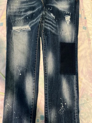 Mörkblå distressed jeans Dsquared2 - Säljer ett par mörkblå jeans från Dsquared2 med slitningar, färgstänk och en stor svart patch på ena benet. Jeansen har en trendig, avslappnad look med unika detaljer och raka ben. Perfekta för dig som gillar streetwear och statement-plagg.
