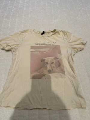 Beige t-shirt med hundmotiv Gina Tricot - Beige t-shirt från Gina Tricot i bomull med korta ärmar och ett tryck framtill av en hund och texten 'Me taking my third 'self care day' in a row, knowing I'm just lazy'. Perfekt för dig som gillar roliga prints och chill vibes.