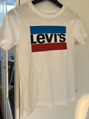 Vit Levi's t-shirt med logga - Säljer en klassisk vit t-shirt från Levi's med stor tryckt logga i blått, svart och rött på bröstet. T-shirten har rund halsringning och korta ärmar. Perfekt till jeans eller shorts för en enkel och snygg look.