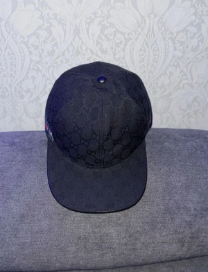 Svart keps från Gucci med GG-mönster - Snygg svart keps från Gucci med klassiskt GG-monogram över hela kepsen. På sidan finns en ikonisk grön och röd rand. Justerbar rem baktill för perfekt passform. Materialet ser ut att vara bomullscanvas med exklusiv känsla.