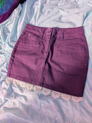 Lila jeanskjol med spetskant - Remake gjord av mig! Supersöt lila kort jeanskjol med vita spetsdetaljer längst ner. Kjolen har klassiska jeansfickor fram och bak, bälteshällor och knappstängning. Perfekt för dig som vill ha en unik twist på din outfit!