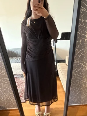 Brun meshklänning med drapering - Snygg brun långklänning i mesh med draperade detaljer och långärmad design. Klänningen har en transparent känsla i ärmarna och ett elegant fall. Perfekt för dig som gillar en stilren och trendig look.