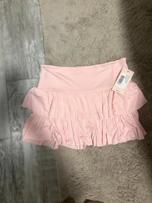 Ljusrosa volang kjol - Supersöt ljusrosa kjol från Nelly med dubbla volanglager och bred midjeresår. Kjoldesign framtill, perfekt för en trendig och bekväm look. Tillverkad i mjukt, stretchigt material som ger en skön känsla hela dagen.