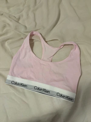 Ljusrosa bh från Calvin Klein - Säljer en ljusrosa bh från Calvin Klein med bred resår och logga runt om. Bh:n har brottarrygg och är tillverkad i mjukt stretchigt material som ger en bekväm passform. Perfekt för dig som gillar enkel och stilren design.