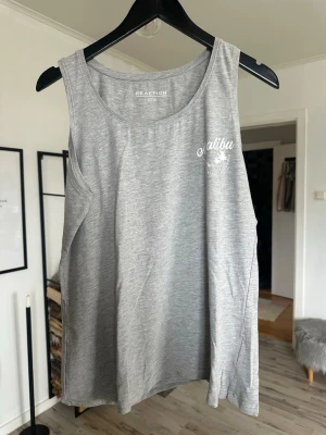 Grå linnetopp från Reaction XXL - Snygg grå ärmlös linnetopp från Reaction med vit tryckt text 'Malibu Palm Springs California' och palmträd på bröstet. Perfekt för en avslappnad stil och enkel att matcha med jeans eller shorts.