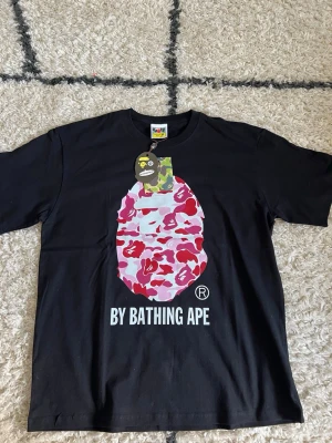 Svart BAPE t-shirt med rosa camo - Säljer en svart t-shirt från A Bathing Ape med ikoniskt rosa och rött camo-mönster på bröstet och stor BAPE-logga. T-shirten har rund hals och korta ärmar. Trycket är riktigt färgstarkt och sticker ut, perfekt för dig som gillar streetwear.