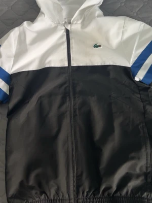Lacoste vindjacka svart/vit/blå - Snygg vindjacka från Lacoste i svart och vitt med blå detaljer på armarna Lacoste . Jackan har luva, dragkedja framtill och elastisk nederkant. Perfekt för en sportig och trendig look. Den är äkta och helt ny pris kan diskuteras och om ni har frågor de ba ställa 