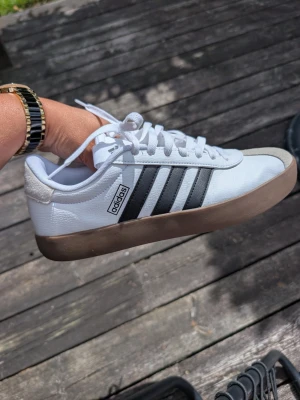 Vita Adidas sneakers med svarta ränder stl 43 1/3 - Snygga vita Adidas sneakers med klassiska svarta ränder på sidan och brun sula. Skorna har lågt skaft, vita skosnören och detaljer i mocka framtill. Perfekta för en clean och sportig look.