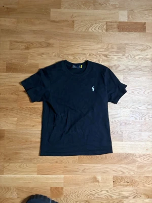 Svart t-shirt från Polo Ralph Lauren - Klassisk svart t-shirt från Polo Ralph Lauren med den ikoniska loggan broderad på bröstet. T-shirten har rund hals och korta ärmar, tillverkad i mjuk bomull för en skön känsla. Perfekt basplagg till garderoben.