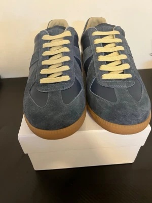 Maison Margiela  - Snygga sneakers i blått med både mocka och skinnpaneler. Skorna har beige snörning och en klassisk gummisula i brunt. Perfekta för dig som gillar stilrena och trendiga sneakers med retrovibbar.Maison Margiela skor väldigt fina och speciella, är du sugen på ett par nya sommar skor är det rätt vall till dig;) kvitto finns!!!!