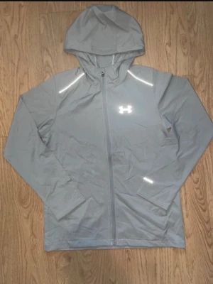 Grå vindjacka från Under Armour - Snygg grå vindjacka från Under Armour med hel dragkedja, huva och reflekterande detaljer på axlar och ärm. Tillverkad i lätt och tunt material som passar perfekt för träning eller promenader. Logga på bröstet och smidig passform.