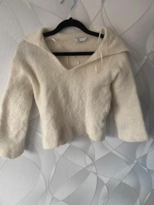 Krämvit stickad tröja med krage - Mysig krämvit stickad tröja med bred krage och v-ringning. Tröjan har långa ärmar och ribbstickade muddar vid nederkant. Perfekt för kalla dagar och enkel att styla till olika outfits. Den har blivit lite mindre i tvätten.