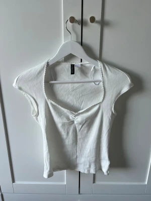Vit ribbad topp med v-ringning - Säljer en vit ribbad topp med v-ringning och korta ärmar. Toppen har en söt liten volangkant vid halsen och är tillverkad i ett mjukt, stretchigt material. Perfekt att matcha med jeans eller kjol för en fräsch look.
