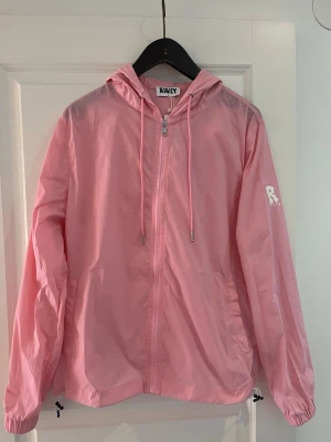 Rosa vindjacka med huva från Risky - Snygg rosa vindjacka från Risky med dragkedja framtill, justerbar huva och resår i ärmsluten. Jackan har ett vitt tryck på ena ärmen och stort vitt tryck på huvan. Perfekt för blåsiga dagar och enkel att matcha till streetwear. Slutsåld I storlek S och kommer inte komma in på lager