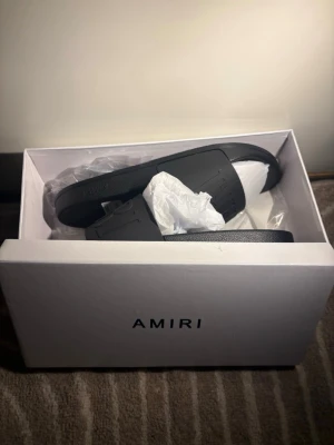 Svarta slides från Amiri - Snygga svarta slides från Amiri med bred rem över foten och märkets logga både på sulan och ovandelen. Tillverkade i slitstarkt gummi för en clean och modern look. Perfekta för sommaren eller att glida runt i hemma.