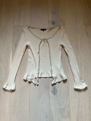 Beige/off white ribbad kofta från Vila - Söt beige ribbad kofta från Vila med långa ärmar och volangdetaljer. Koftan har öppen front med knytband. Perfekt för lager på lager! Lite nopprig men inga tydliga defekter