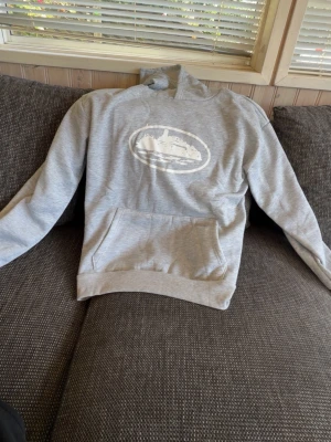 cortiez hoodie - Säljer en ljusgrå hoodie med huva och magficka. På bröstet finns ett vitt tryck med motiv i oval form. Hoodien har ribbade muddar vid ärmslut och nederkant. Perfekt för en avslappnad och chill stil.