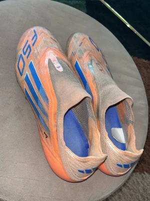 Orange elite fotbollsskor utan snörning - Säljer ett par snabba fotbollsskor i orange med blå detaljer och strömlinjeformad design. Skorna har en sockliknande öppning utan snörning och är tillverkade i syntetmaterial för lätt vikt och bra bollkontroll. Perfekta för dig som vill sticka ut på planen.