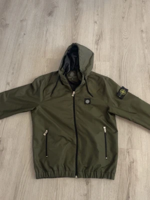 Grön vindjacka med huva Stone Island - Snygg olivgrön vindjacka från Stone Island med huva och dragkedja framtill. Jackan har två fickor med dragkedja, elastisk nederkant och ärmslut samt den klassiska logotypen på vänster ärm. Perfekt för blåsiga dagar. Skulle säga passar M och L