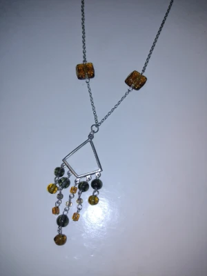 Vintage ilverfärgat halsband med bärnstensfärgade glaspärlor/kristaller - Unikt halsband med silverfärgad kedja, två fyrkantiga bärnstensfärgade "glaspärlor" och ett diamantformat hänge. Från hänget hänger flera små glaspärlor i bärnstensfärg, grönt/gult och grått. Vet tyvärr inte vad det är för typ av metall eller säkert vad pärlorna är gjorda i för material, men tror glas/kristall. Kedjan är ca 45 + 7 cm lång och hänget ca 8 cm.