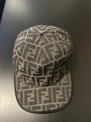 Grå keps med Fendi-logga - Säljer en grå keps från Fendi med det klassiska FF-monogrammet över hela kepsen. Kepsen har böjd skärm och är tillverkad i ett tygmaterial med exklusiv känsla. Perfekt för dig som vill ha en statement-accessoar med lyxig touch.