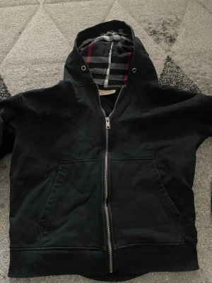 Svart hoodie med dragkedja från Burberry - Säljer en svart hoodie från Burberry med dragkedja framtill och klassiskt rutigt foder i huvan. Hoodien har två stora fickor fram och metallöglor vid huvan. Perfekt för dig som gillar stilrena och exklusiva plagg.