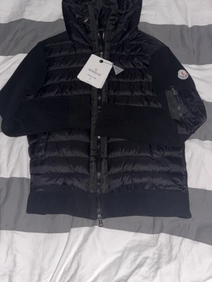 Svart monclear cardigan  - Snygg svart cardigan från Moncler med stickade ärmar och huva. Jackan har dragkedja framtill, Moncler-logga på ärmen och en ficka med dragkedja på ena ärmen. Perfekt för kalla vinterdagar och riktigt trendig look. Moncler märket går att scanna och du kommer direkt till monclears hemsida 