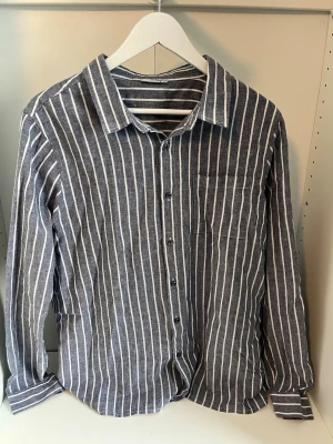 Randig linneskjorta från Marinel - Snygg blå och vit randig skjorta från Marinel i luftig linnemix. Skjortan har klassisk krage, bröstficka och knäppning framtill. Perfekt för en avslappnad och stilren look.