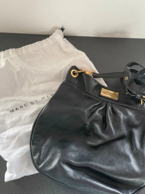 Svart handväska Marc by Marc Jacobs - Snygg svart handväska i mjukt läder från Marc by Marc Jacobs. Väskan har guldfärgade metalldetaljer, justerbar axelrem och en dekorativ metallbricka framtill. Medföljer original dustbag. Perfekt för dig som gillar klassisk och stilren design. Dustbag medföljer.  Bredd 29 cm, höjd 31 cm, djup ca 7 cm  Skicka meddelande för fler bilder💞💞