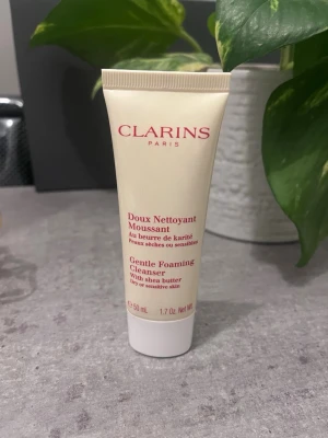 Clarins Gentle Foaming Cleanser 50ml - Clarins Gentle Foaming Cleanser med sheasmör är en mild rengöringskräm för torr eller känslig hud. Kommer i en beige tub med röd text och rymmer 50 ml. Perfekt för daglig ansiktsrengöring och lämnar huden mjuk och fräsch.