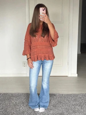 Rostfärgad virkad stickad tröja - Säljer en snygg rostfärgad stickad tröja med virkat mönster och volangkant. Tröjan har trekvartsärmar och en lös, avslappnad passform. Perfekt att styla med jeans för en trendig look.