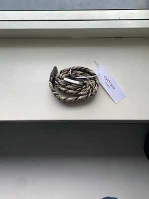 Randigt flätat bälte från Onewear - Snyggt randigt bälte från Onewear i beige och brunt. Bältet är flätat och har ett metallspänne samt detaljer i brunt. är helt nytt! Skriv vid dom minsta frågorna 
