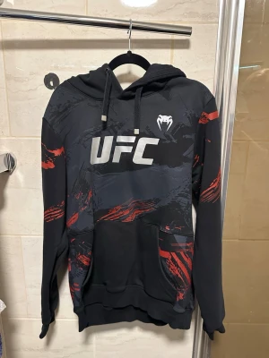 Svart och röd UFC hoodie - Säljer en svart hoodie med UFC-tryck på bröstet och coola röda och grå grafiska detaljer. Hoodien har huva med dragsko och en stor magficka. Perfekt för dig som gillar sportig streetwear och vill sticka ut lite extra.