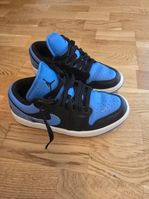 Nike Air Jordan 1 Low Blue/Black - Säljer ett par Nike Air Jordan 1 Low sneakers i blått och svart med vita sulor. Skorna har klassisk låg siluett, svart swoosh och svarta skosnören. Ovandelen är i läder med perforerad tåbox för extra ventilation. Perfekta för dig som gillar streetwear och snygg design.