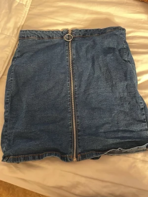 Blå jeanskjol med dragkedja - Snygg blå jeanskjol i kort modell med en stor silverfärgad dragkedja framtill. Kjolen har en enkel och klassisk design som passar till det mesta. Perfekt för dig som gillar denim och vill ha en trendig look.