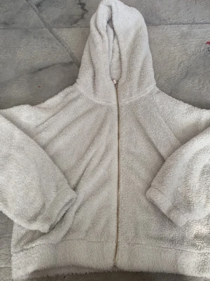 Mysig beige fleecehoodie med dragkedja - Supermjuk och fluffig beige hoodie i fleece med dragkedja framtill och stor huva. Perfekt för chill hemma eller när du vill vara extra bekväm. Vida ärmar och avslappnad passform gör den extra cozy.