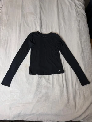 Svart långärmad topp Hollister - En svart långärmad topp från Hollister med rund halsringning och diskret logga broderad vid nederkanten. Modellen är slim fit och passar perfekt till jeans eller kjol. Enkel och stilren design som funkar till det mesta.