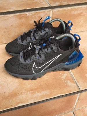 Nike react vision - Snygga svarta Nike sneakers med grå och blå detaljer, vit swoosh på sidan och grov sula. Ovandelen är i mesh och syntetmaterial, med coola blå och silvriga detaljer vid hälen. Perfekta för dig som gillar sportig och modern stil.